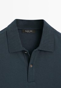 Ciemnozielona koszulka polo wykonana z teksturowanej tkaniny. Posiada klasyczny kołnierz, listwę z trzema guzikami oraz metkę wszytą wewnątrz kołnierza.