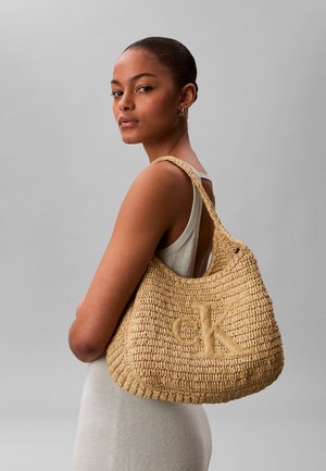 Young woman with pulled-back hair wearing a sleeveless dress and carrying a large woven beige bag with "ck" logo on shoulder.