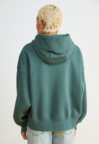 Πράσινη υπερμεγέθη φούτερ με μεγάλη κουκούλα. Διαθέτει λεία υφή και ribbed μανσέτες. Φοριέται πάνω από ανοιχτό μπλε τζιν με λεκέδες.