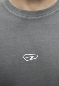 Κοντινό ενός γκρι μπλουζάκι με στρογγυλή λαιμόκοψη και μικρό λευκό γεωμετρικό λογότυπο στο κέντρο του στήθους.