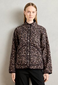 Woman wearing a black quilted jacket with small beige floral patterns and silver snap buttons, paired with black pants against a plain background.