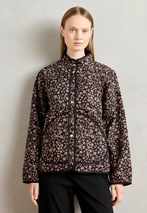 Kvinne iført en svart quiltet jakke med små beige blomster mønster og sølvfargede trykknapper, kombinert med svarte bukser mot en ensfarget bakgrunn.