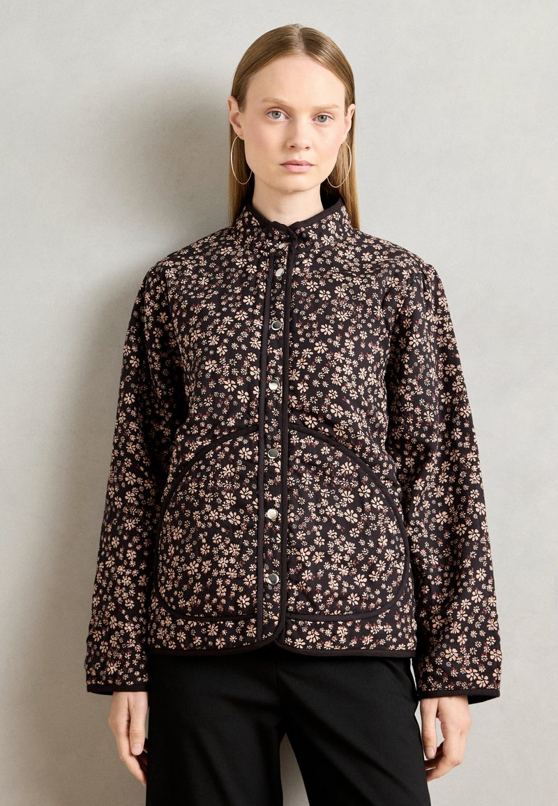 Woman wearing a black quilted jacket with small beige floral patterns and silver snap buttons, paired with black pants against a plain background.