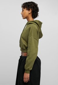 Olive green cropped hoodie with a hood, long sleeves, and a front zipper. Paired with black, loose-fitting pants, showcasing soft fabric.