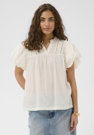 Kobieta o kręconych brązowych włosach ubrana w luźną białą bluzkę z koronkowymi detalami i bufkami, w połączeniu z jasnoniebieską spódnicą dżinsową i złotą bransoletką.