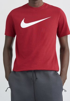 Mand iført en rød Nike t-shirt med stort hvidt swoosh-logo, kombineret med mørkegrå bukser med snøre, hænder i lommerne.