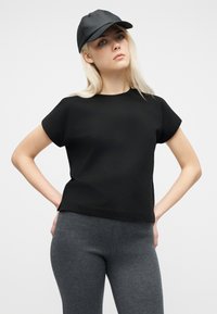 Γυναίκα που φοράει μαύρο καπέλο και κοντομάνικη μαύρη μπλούζα με σκούρο γκρι κολάν, στέκεται με τα χέρια στη μέση μπροστά σε απλό φόντο.