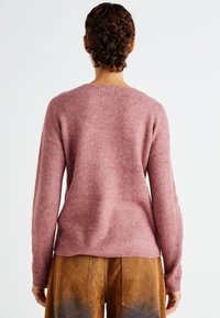 Helle rosa gerippte Strickjacke mit lockerer Passform, langen Ärmeln und weitem Ausschnitt, kombiniert mit braunen Wildlederhosen.