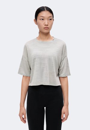 Femme aux cheveux noirs et raides attachés en arrière, portant un haut ample gris clair à manches courtes et un pantalon noir ajusté sur un fond uni.