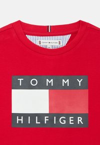 Punainen puuvillainen t-paita, jossa keskellä oleva grafiikka, jossa lukee valkoisin kirjaimin "TOMMY" ja "HILFIGER" mustalla pohjalla ja monivärisillä suorakaiteen muotoisilla korostuksilla.