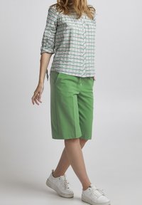 Žena v zelených šortkách po kolena, bílých teniskách a bílé košili se zelenými pruhy, stojící na pozadí světle jednobarevného pozadí.