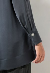 Camicetta grigio scuro con una texture liscia; presenta un bottone con un motivo floreale al polsino e una silhouette dritta.