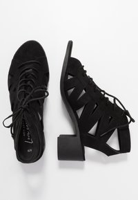 Czarne zamszowe buty na obcasie z wyciętym wzorem, sznurowanym przodem i szerokim obcasem, z teksturowanymi akcentami i solidną podeszwą.