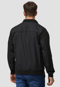 Black bomber jacket with a collar, relaxed fit, ribbed cuffs and waistband, and a smooth, matte texture. Worn over blue jeans.
