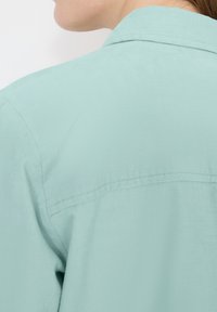 Gros plan sur l'épaule et le cou d'une personne portant une chemise à col bleu clair sur un fond uni.