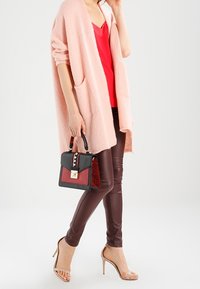 Light pink oversized cardigan with pockets, red v-neck top, brown fitted pants, and a black handbag with red accents and decorative hardware.
