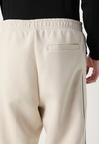 Lys beige sweatpants med elastisk talje, featuring en enkelt baglomme og en slank side stripe i sort. Glat tekstur.