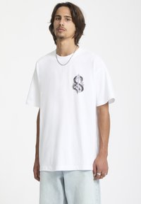 T-shirt oversize en coton blanc avec un design graphique en violet et noir sur la poitrine. Manches courtes et encolure ronde.