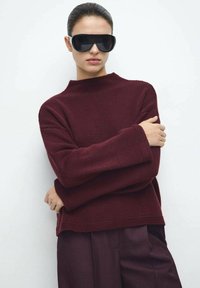 Maglione bordeaux a maglia con collo alto, texture a coste e maniche larghe, abbinato a pantaloni scuri e occhiali da sole neri oversize.