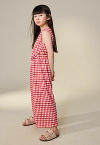 Pige med langt mørkt hår iført ærmeløs rød-hvid ternet kjole og beige sandaler med spænde, stående i profil mod ensfarvet baggrund.