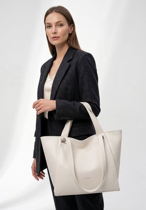 Vrouw met een zwarte blazer houdt een grote witte leren draagtas met twee banden tegen een effen achtergrond.