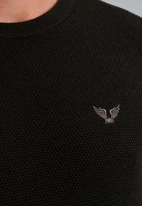 Zwarte gebreide trui met een ribbelkraag en een klein geborduurd logo met vleugels in het grijs op de linkerborst.