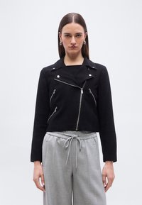 Γυναίκα με ίσια μαλλιά που φοράει ασημένια σκουλαρίκια κρίκους, μαύρο μπουφάν με φερμουάρ και χαλαρό γκρι παντελόνι με κορδόνι, μπροστά σε απλό λευκό φόντο.