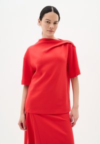 Röd kortärmad topp gjord av mjukt tyg; har en draperad design på ena axeln och en avslappnad passform, i kombination med matchande underdel.
