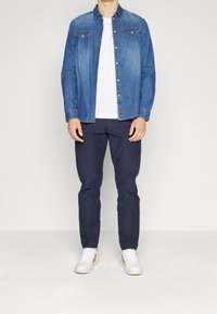 Camisa de mezclilla en azul claro con botones a presión y yugo en punta, combinada con pantalones oscuros de color marino y zapatillas blancas con acentos rojos.