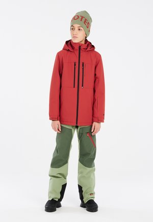 Giacca impermeabile rossa con cappuccio e tasche con zip, abbinata a pantaloni da sci verdi con accenti neri a contrasto e una tasca laterale con zip.