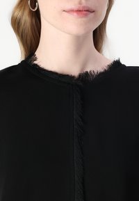 Svart topp med rund halslinning og frynse detaljer langs kanten, glatt stofftekstur og minimalistisk design.