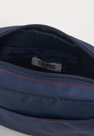 Granatowa saszetka z czerwonym przeszyciem, częściowo otwarty suwak, ukazujący wewnętrzną metkę materiałową z napisem Tommy Jeans.