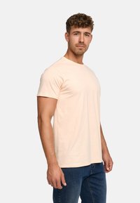 T-shirt à manches courtes de couleur pêche clair en coton doux. Présente un col rond et est associé à un jean bleu foncé. Design minimaliste.