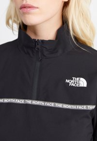 Veste noire à zip quart avec un col montant. Présente un logo blanc sur la poitrine et un graphique du nom de la marque répétitif sur le devant.