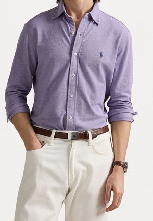 Uomo che indossa una camicia viola chiaro con bottoni e maniche arrotolate, pantaloni bianchi, cintura di pelle marrone e orologio da polso marrone.