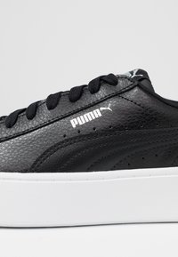 Crne sintetičke kožne tenisice s teksturiranim gornjištem, bijelim gumenim đonom i crnim vezicama. Sadrži PUMA logotip u bijeloj boji.