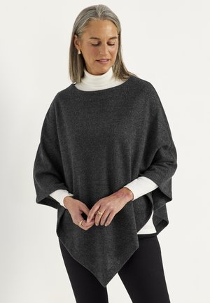 Woman with gray hair wearing gold earrings, a dark gray poncho over a white turtleneck and black pants, standing against a light background.