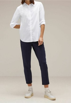 Personne portant une chemise blanche à manches longues boutonnée, un pantalon court bleu marine, des chaussettes blanches et des baskets blanches, debout devant un fond uni.