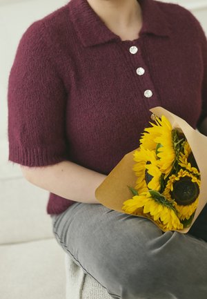 Persoon met een donkerrode gebreide top met grote knopen houdt een bos heldergele zonnebloemen vast, verpakt in bruin papier.