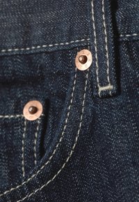 Σκούρο μπλε τζιν ύφασμα με χαλκούς τρούκς και αντίθετη μπεζ ραφή. Κοντινό πλάνο λεπτομέρειας τσέπης και υφής υφάσματος.