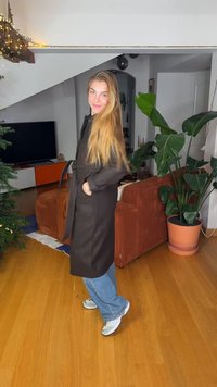 Lange donkerbruine jas met een ceintuur, voorzien van zijzakken. Gedragen over een blauwe spijkerbroek en witte sportschoenen, gesitueerd in een huiselijke omgeving.