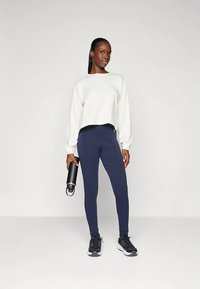 Ljusgrå crop-top sweatshirt och marinblå leggings i stretchtyg. Svarta sneakers och en svart vattenflaska i handen.