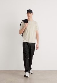 Ανοιχτό μπεζ μπλουζάκι με μικρό λογότυπο, μαύρο παντελόνι με υποτονική υφή και άσπρα αθλητικά παπούτσια. Η φιγούρα κρατά μια μαύρη ζακέτα στον έναν ώμο.