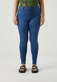 Jeans skinny azules con cintura alta y presillas para cinturón, que cuentan con dos bolsillos frontales y una textura de denim suave. Llevados con sandalias negras.