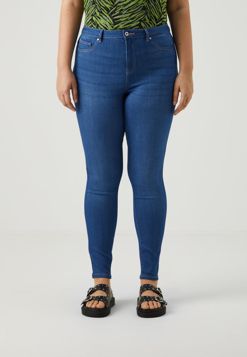Jeans skinny azules con cintura alta y presillas para cinturón, que cuentan con dos bolsillos frontales y una textura de denim suave. Llevados con sandalias negras.