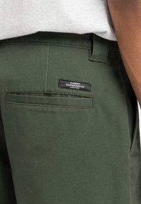 Pantalons chino vert olive avec une finition texturée, dotés d'une poche arrière avec une étiquette noire portant les mots "ELEMENT SKATEBOARD CO."