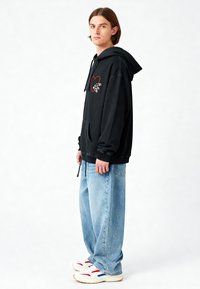 Felpa nera con grafica di un cuore rosso e dettagli bianchi, abbinata a jeans larghi blu chiaro e sneaker bianche con accenti rossi e blu.