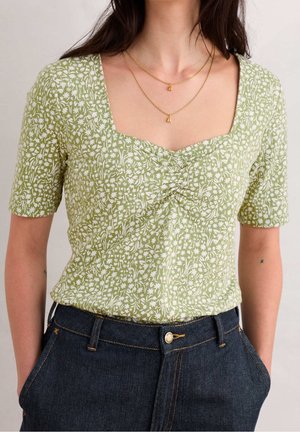 Femme portant un haut vert à manches courtes avec un motif floral blanc et un col froncé, associé à un jean en denim bleu foncé et des colliers dorés superposés.