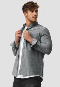 Camicia grigia con bottoni e maniche lunghe, tessuto morbido, tasca singola sul petto, indossata sopra una t-shirt bianca da una modella su sfondo neutro.