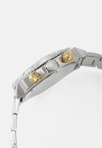 Metallklokke med sølvarmbånd, med rund klokkehus, gull- og sølvkrone, og en strukturert bezel. Klart detaljer på klokkens skjerm.